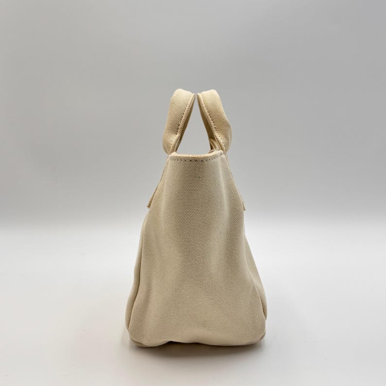 May include: A small, beige canvas tote bag with two sturdy handles. The bag has a simple, rounded shape and a textured surface. The handles are attached at the top of the bag, and the bag appears to be empty.