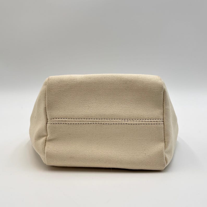 May include: A beige canvas tote bag, viewed from the bottom. The bag has a rectangular shape with a stitched seam detail across the base. The fabric appears to be a sturdy, woven material, suitable for carrying items.