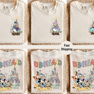 以下が含まれることがあります： ディズニーキャラクターのグラフィックが施されたクリーム色のTシャツ。上段には「Oliver」、「Olivia」、「James」と名前が入ったシャツがあります。下段には、ミッキーマウスと仲間たちが描かれた「Disneyland 1955」のデザインのシャツが表示されています。