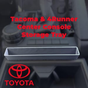 May include: A black rectangular center console storage tray with a silver trim. The text "Tacoma & 4Runner Center Console Storage Tray" is in red. The Toyota logo and the word "TOYOTA" are also in red.