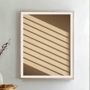 May include: A framed artwork with a beige background and diagonal shadow lines. The artwork is in a light wood frame with a white border. A wooden bowl, vase, and woven basket are in the foreground.