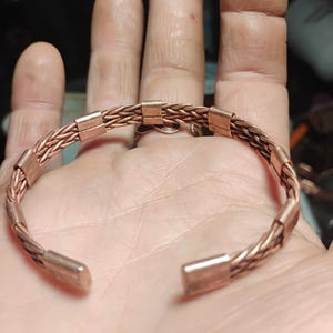 May include: A copper-coloured bracelet with a braided design. The bracelet has a cuff style with rectangular end caps and rectangular accents along the braided section. The bracelet is shown on a hand.