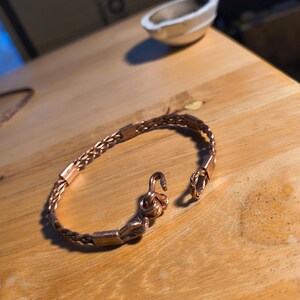 May include: A copper bracelet with a woven chain pattern and a hook clasp. The bracelet is displayed on a wooden surface. The clasp is open, showcasing the detailed metalwork.