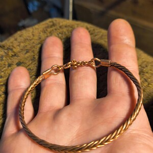 May include: A gold-coloured bracelet with a braided design. The bracelet is made of thin, twisted metal strands, creating a textured appearance. The clasp and connecting elements are also gold-coloured. The bracelet is a simple, elegant piece of jewellery.