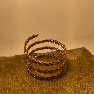 May include: A copper-colored, spiral-shaped bracelet. The bracelet is made of twisted wire and rests on a textured, golden-brown surface. The spiral design creates a unique, eye-catching accessory.