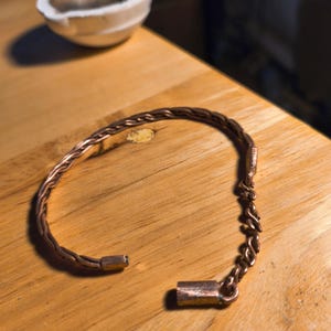 May include: A copper-coloured bracelet with a braided design and a chain closure. The bracelet is open, revealing the clasp. A small, white bowl sits in the background on a wooden surface.