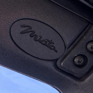 May include: Close-up of a black interior car panel featuring an oval emblem with the cursive word "Miata". The emblem is centered on the panel, with a textured surface. Two screws are visible on the right side of the panel.