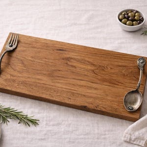May include: A rectangular wooden serving board with a fork and spoon attached. The board is light brown and has a rustic appearance. A small bowl of olives, bread slices, and rosemary sprigs are nearby.