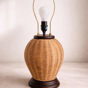 May include: A decorative table lamp with a woven wicker shade and a dark brown wooden base. The lamp has a brass-colored metal frame and a white light bulb. The lamp is designed for indoor use.