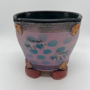 Peut inclure: Pot en céramique fait à la main avec un extérieur lavande, orné de pois turquoise et de petits ornements de couleur beige. Le pot repose sur trois pieds rouges rectangulaires. Le bord est noir.