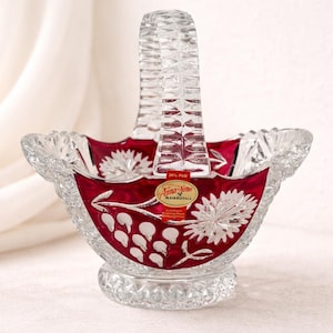 May include: A clear crystal basket with a red interior and a handle. The basket features a floral design and a label that reads "Anna Hütte Bleikristall". The basket is approximately 6 inches tall.