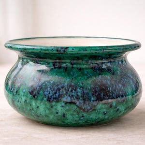 May include: A handcrafted ceramic bowl with a glossy, speckled glaze in shades of green and blue. The bowl has a rounded shape with a slightly flared rim and a white interior. This decorative bowl is suitable for home decor.