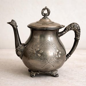 May include: Antique silver teapot with ornate floral engravings. The teapot features a curved spout, a decorative handle, and a lid with a loop finial. The body of the teapot rests on three small feet. A classic piece for tea lovers.