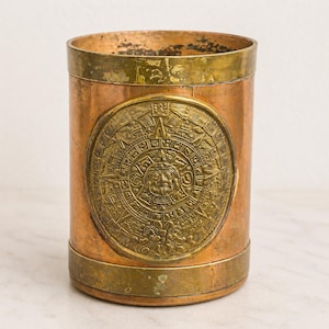 May include: Cylindrical copper container with brass accents. A large, circular brass emblem, possibly a calendar or sun symbol, is affixed to the front. The container has a vintage, aged appearance, with visible wear and patina on the metal surfaces.