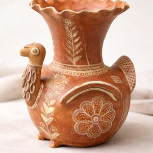 May include: A handcrafted, terracotta-colored ceramic vase with a bird-like design. The vase features a ruffled rim, a bird head, and tail, and is adorned with floral and leaf patterns in a lighter hue. The vase is approximately 8 inches tall.