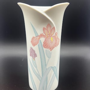 Puede incluir: Un jarrón de cerámica blanco con la parte superior acampanada, con un diseño floral de iris rosas y amarillos con hojas verde azulado. El jarrón tiene un acabado liso y brillante y una forma alta y esbelta.