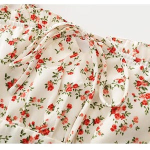Puede incluir: Primer plano de una tela color crema con un estampado floral de peque&ntilde;as rosas rojas y hojas verdes. La tela est&aacute; fruncida en la parte superior y atada con cintas color crema, posiblemente una prenda.