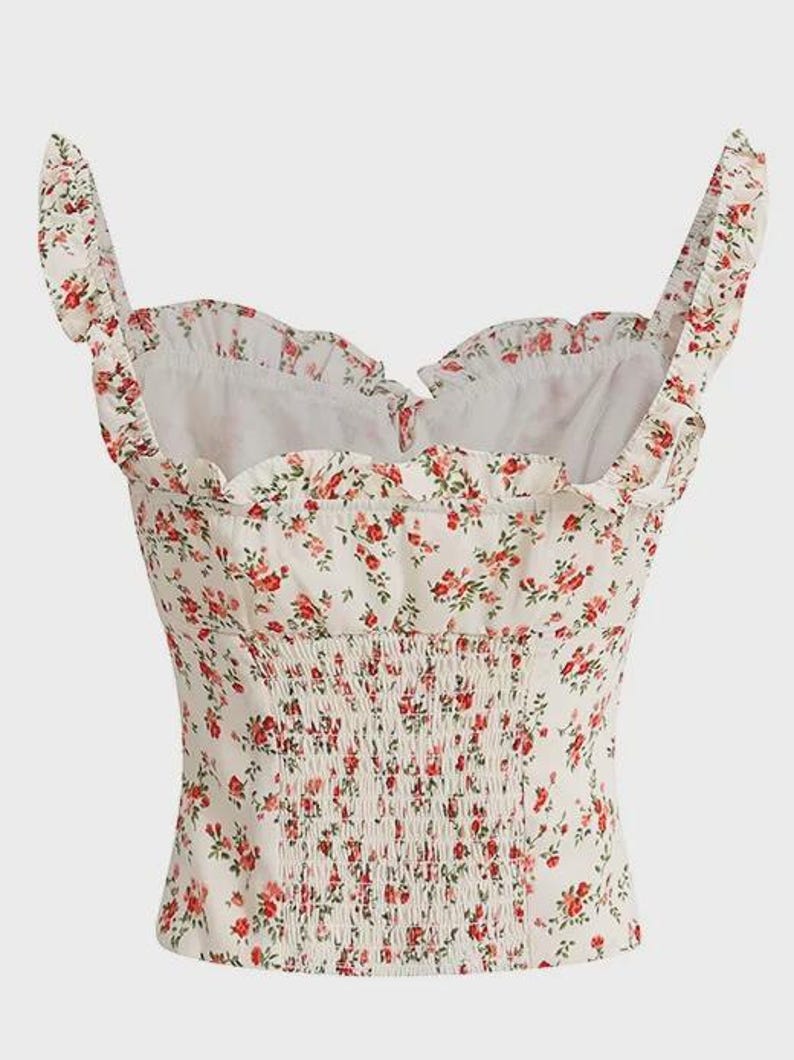 Puede incluir: Top blanco con estampado floral de flores rojas y hojas verdes. Presenta un escote coraz&oacute;n, ribetes con volantes y un corpi&ntilde;o fruncido. Las correas tambi&eacute;n est&aacute;n adornadas con volantes. Un estilo femenino y rom&aacute;ntico.