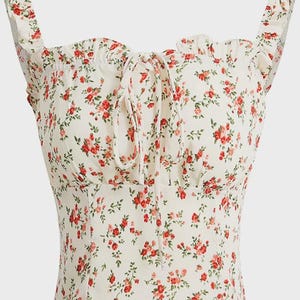 Puede incluir: Una blusa floral color crema con rosas rojas y hojas verdes. La blusa presenta un escote cuadrado, tirantes con volantes y un detalle de lazo en el busto. La tela tiene una apariencia delicada y ligera, adecuada para climas c&aacute;lidos.