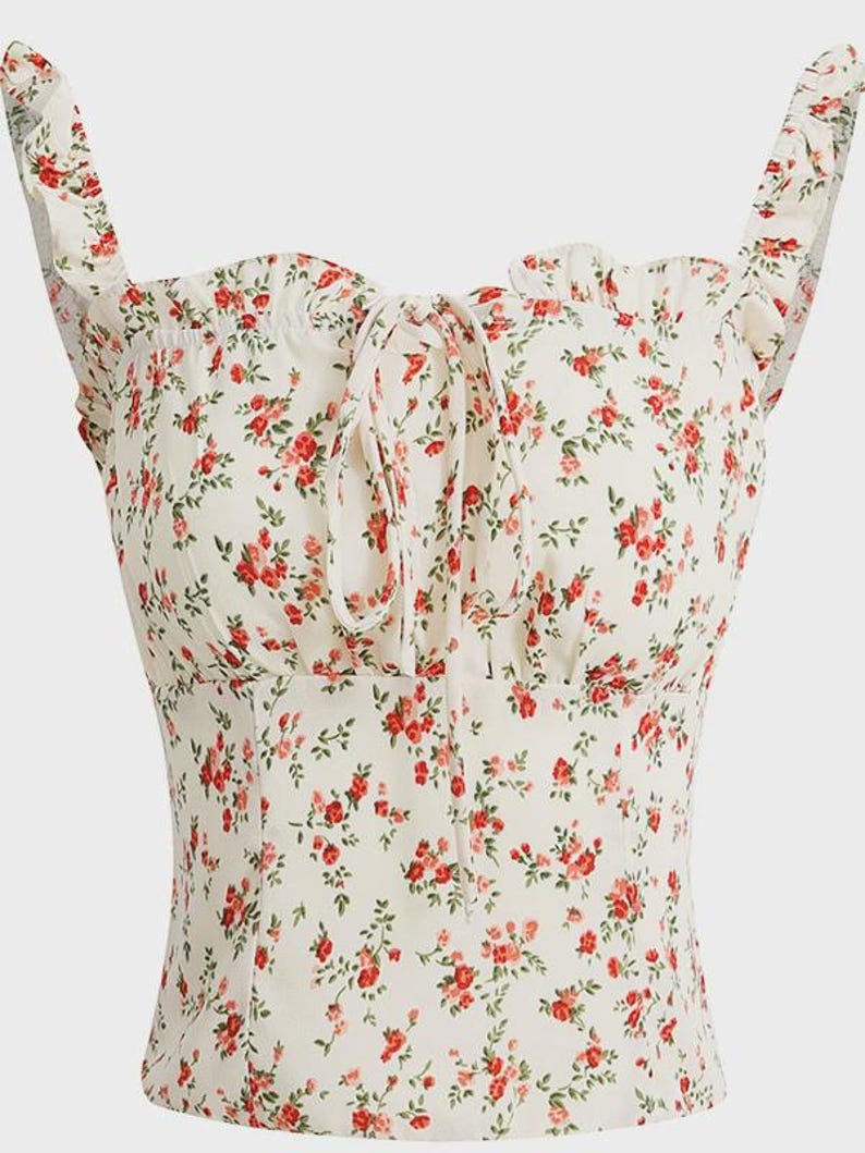 Puede incluir: Una blusa floral color crema con rosas rojas y hojas verdes. La blusa presenta un escote cuadrado, tirantes con volantes y un detalle de lazo en el busto. La tela tiene una apariencia delicada y ligera, adecuada para climas c&aacute;lidos.