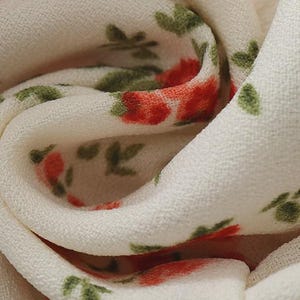 Puede incluir: Primer plano de una tela color crema con un estampado floral. Las flores son rojas con hojas verdes, creando un dise&ntilde;o delicado. La tela es suave y ligera, con una textura visible. El patr&oacute;n se repite.