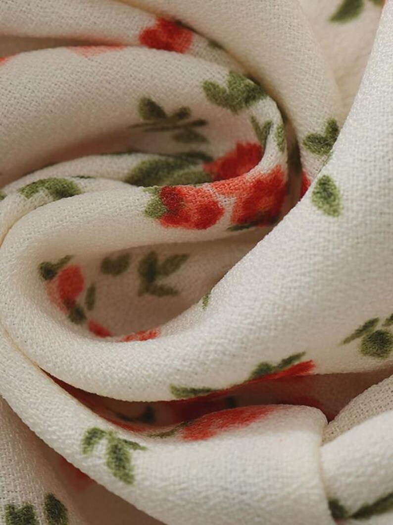 Puede incluir: Primer plano de una tela color crema con un estampado floral. Las flores son rojas con hojas verdes, creando un dise&ntilde;o delicado. La tela es suave y ligera, con una textura visible. El patr&oacute;n se repite.