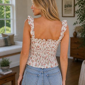 Puede incluir: Top blanco con estampado floral, tirantes con volantes y corpi&ntilde;o fruncido. El top se combina con vaqueros azules de lavado claro. El estampado floral presenta flores rojas y rosas sobre un fondo blanco. El top es de estilo ajustado.