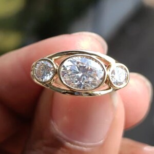May include: A gold ring featuring three clear, round-cut gemstones. The central stone is oval-shaped, flanked by two smaller, round stones. The ring has a delicate, openwork design, and the gemstones sparkle brightly.