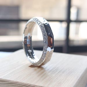May include: A silver-toned ring featuring a faceted outer band and a channel set with a row of small, clear, sparkling stones along the side. The metal surface is highly reflective, showcasing a polished finish and a geometric, multi-faceted design.