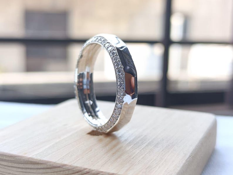 May include: A silver-toned ring featuring a faceted outer band and a channel set with a row of small, clear, sparkling stones along the side. The metal surface is highly reflective, showcasing a polished finish and a geometric, multi-faceted design.