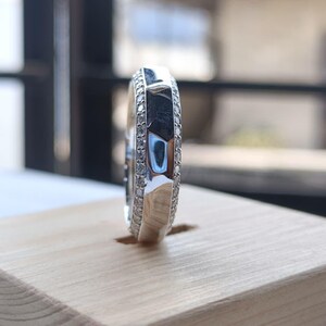 May include: A silver-toned ring featuring a central band with a faceted, reflective surface. The ring is flanked on both sides by two thin rows of small, clear, sparkling stones that encircle the entire circumference of the metal band.