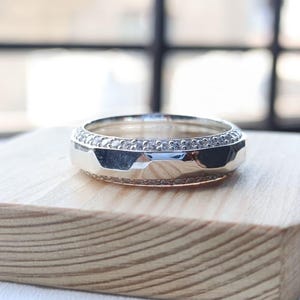 May include: A silver-toned metal ring featuring a central band with a faceted, geometric surface. The ring is bordered on both the top and bottom edges by a thin, continuous row of small, clear, sparkling stones set into the metal band.