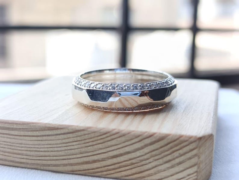May include: A silver-toned metal ring featuring a central band with a faceted, geometric surface. The ring is bordered on both the top and bottom edges by a thin, continuous row of small, clear, sparkling stones set into the metal band.