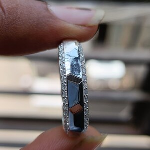 May include: A silver-toned ring featuring a central band with a faceted, geometric surface design. The center section is flanked on both sides by two rows of small, clear, round-cut stones embedded along the edges of the metallic band.