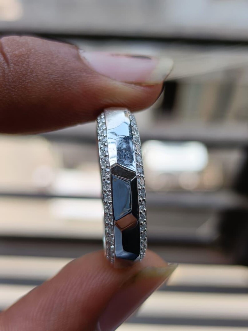 May include: A silver-toned ring featuring a central band with a faceted, geometric surface design. The center section is flanked on both sides by two rows of small, clear, round-cut stones embedded along the edges of the metallic band.