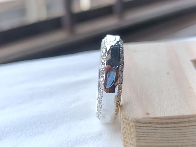 May include: A silver-toned ring featuring a central band with a smooth, reflective surface flanked by two outer rows of small, clear, round-cut stones. The ring is positioned vertically, showcasing its profile and the detailed stone setting.