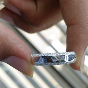 May include: A silver-toned band ring featuring a central row of faceted, geometric metal segments flanked by two thin rows of small, clear, sparkling stones embedded along the top and bottom edges of the band's outer circumference.