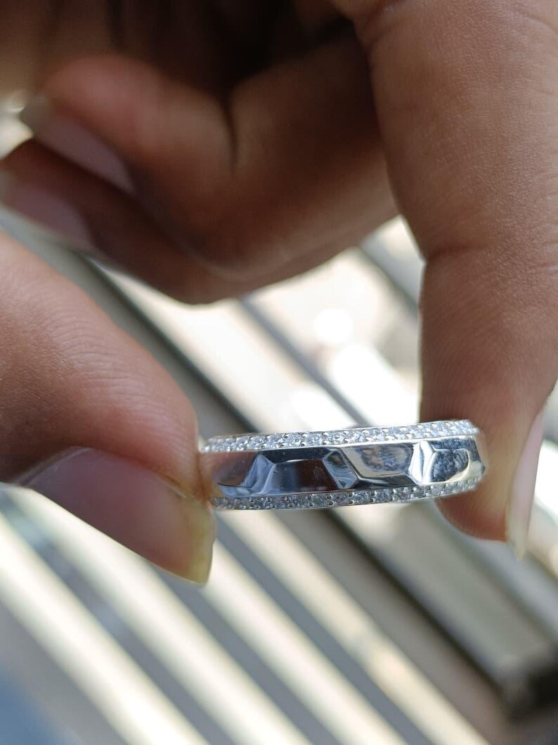 May include: A silver-toned band ring featuring a central row of faceted, geometric metal segments flanked by two thin rows of small, clear, sparkling stones embedded along the top and bottom edges of the band's outer circumference.