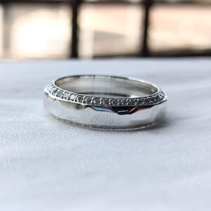 May include: A silver-toned ring featuring a smooth, polished central band flanked by two thin rows of small, clear, sparkling stones. The metal surface reflects light, highlighting the contrast between the reflective center and the textured edges.