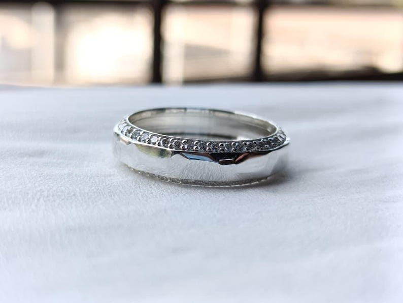May include: A silver-toned ring featuring a smooth, polished central band flanked by two thin rows of small, clear, sparkling stones. The metal surface reflects light, highlighting the contrast between the reflective center and the textured edges.