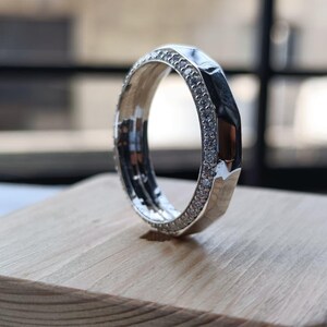 May include: A silver-toned ring featuring a faceted outer edge and a band encrusted with small, clear, sparkling stones along the side. The ring has a polished metallic finish and a distinct geometric design with multiple flat surfaces.