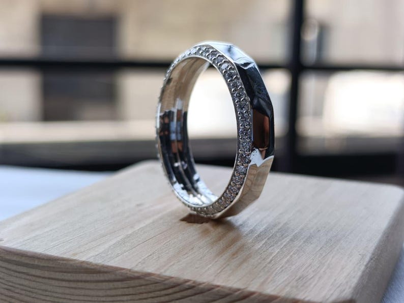 May include: A silver-toned ring featuring a faceted outer edge and a band encrusted with small, clear, sparkling stones along the side. The ring has a polished metallic finish and a distinct geometric design with multiple flat surfaces.