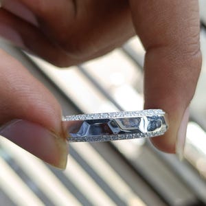 May include: A silver-toned ring featuring a central band with a faceted, geometric surface texture. The ring is bordered on both the top and bottom edges by thin rows of small, clear, sparkling stones that encircle the entire circumference.