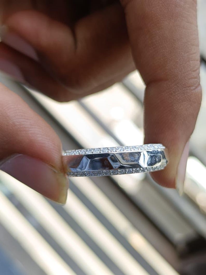 May include: A silver-toned ring featuring a central band with a faceted, geometric surface texture. The ring is bordered on both the top and bottom edges by thin rows of small, clear, sparkling stones that encircle the entire circumference.