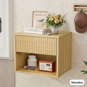 May include: A light wood-toned floating nightstand with a woven drawer front. The nightstand has an open shelf with a small radio, sunglasses, and a decorative vase. A framed print, books, and flowers sit on top. A brown baseball cap hangs on the wall.