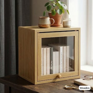 May include: A light oak wooden cabinet with a glass door, displaying books. A small potted plant, a mug, and a candle sit on top. A magnifying glass and glasses are on the table. The word "Wooden" is in the bottom right corner.