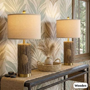 May include: Two wooden table lamps with beige fabric shades. The lamp bases are designed to resemble tree trunks, with a gold-colored base and accents. The lamps are on a dark table with a woven runner, and a decorative basket.