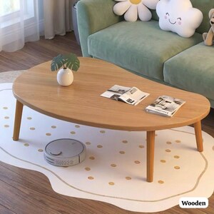 May include: A light brown, oval-shaped wooden coffee table with four angled legs. The table has a smooth surface and is surrounded by a white rug with gold polka dots. A small white vase with green leaves and two magazines are on the table.