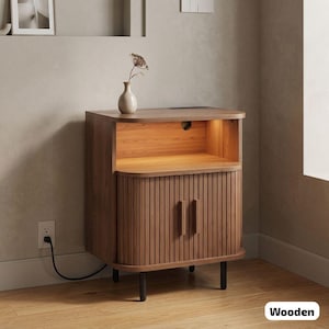 May include: A wooden nightstand with a warm brown finish. The cabinet features a curved front with vertical grooves and two doors. The top has a built-in light and a small vase with a dried flower. The nightstand is supported by black legs.