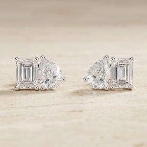 May include: A pair of diamond stud earrings. Each earring features an emerald-cut diamond paired with a pear-shaped diamond. The diamonds are set in a silver or white gold setting. The earrings are displayed on a neutral background.